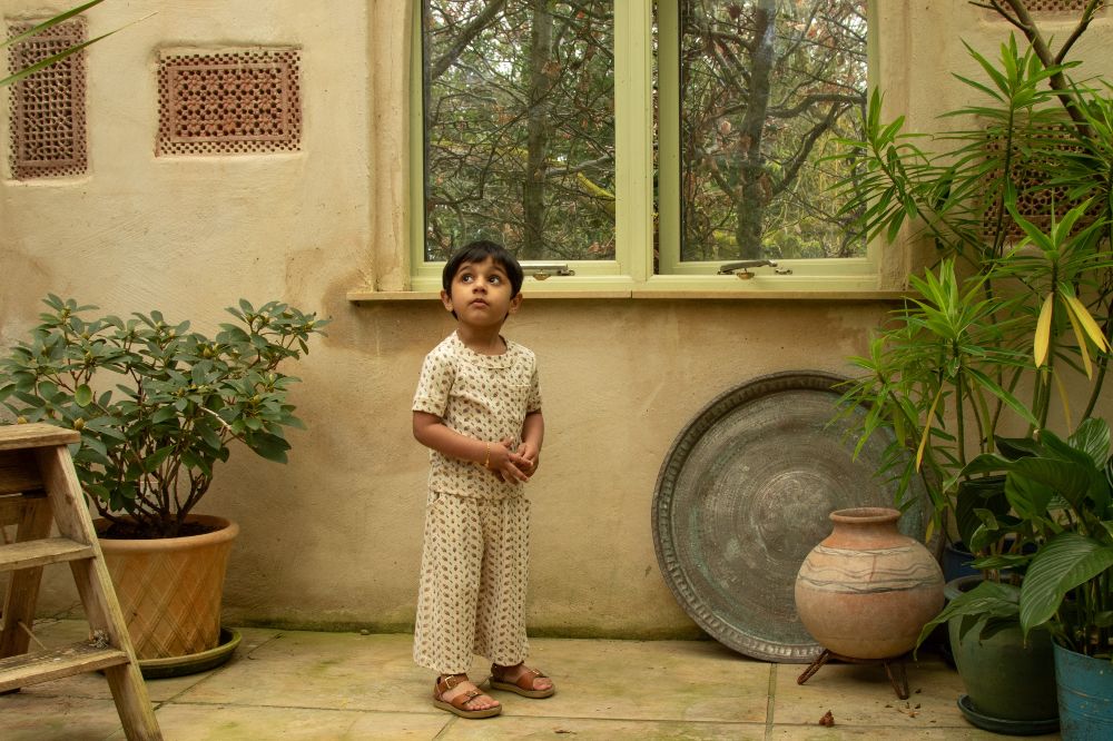 A young boy stood in room full of plants wearing trousers and a top by the children's brand Kari Sako