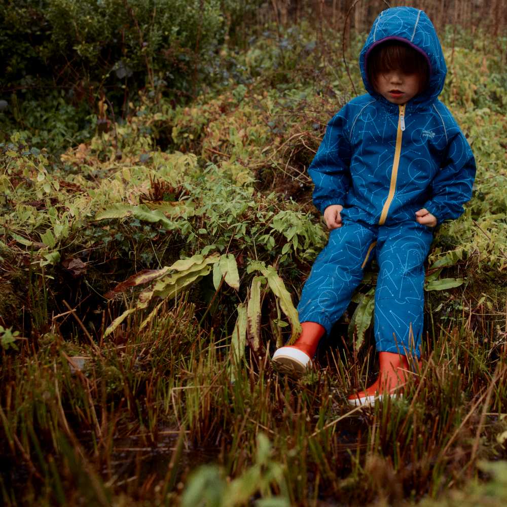 A young child sat on an embankment in a rainsuit from Muddy Puddles Intergalactic Collection