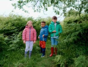 Three children stood outside among trees and bushes wearing outerwear from the Muddy Puddles Intergalactic Collection