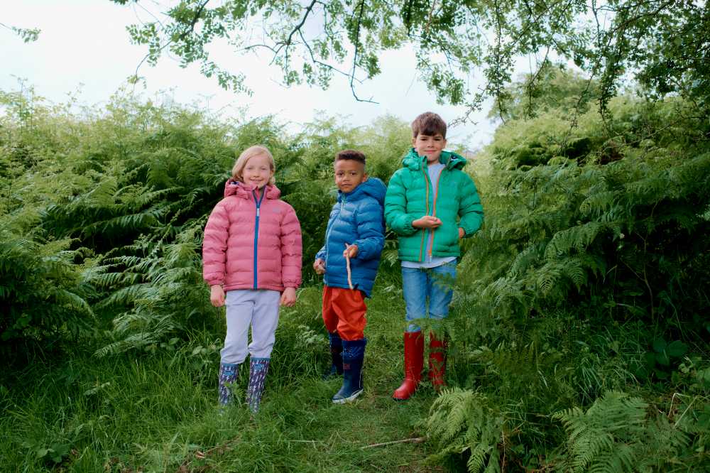 Three children stood outside among trees and bushes wearing outerwear from the Muddy Puddles Intergalactic Collection