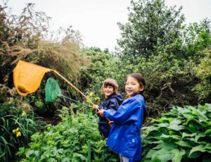 Two children outside in Muddy Puddles rainwear holding fishing nets