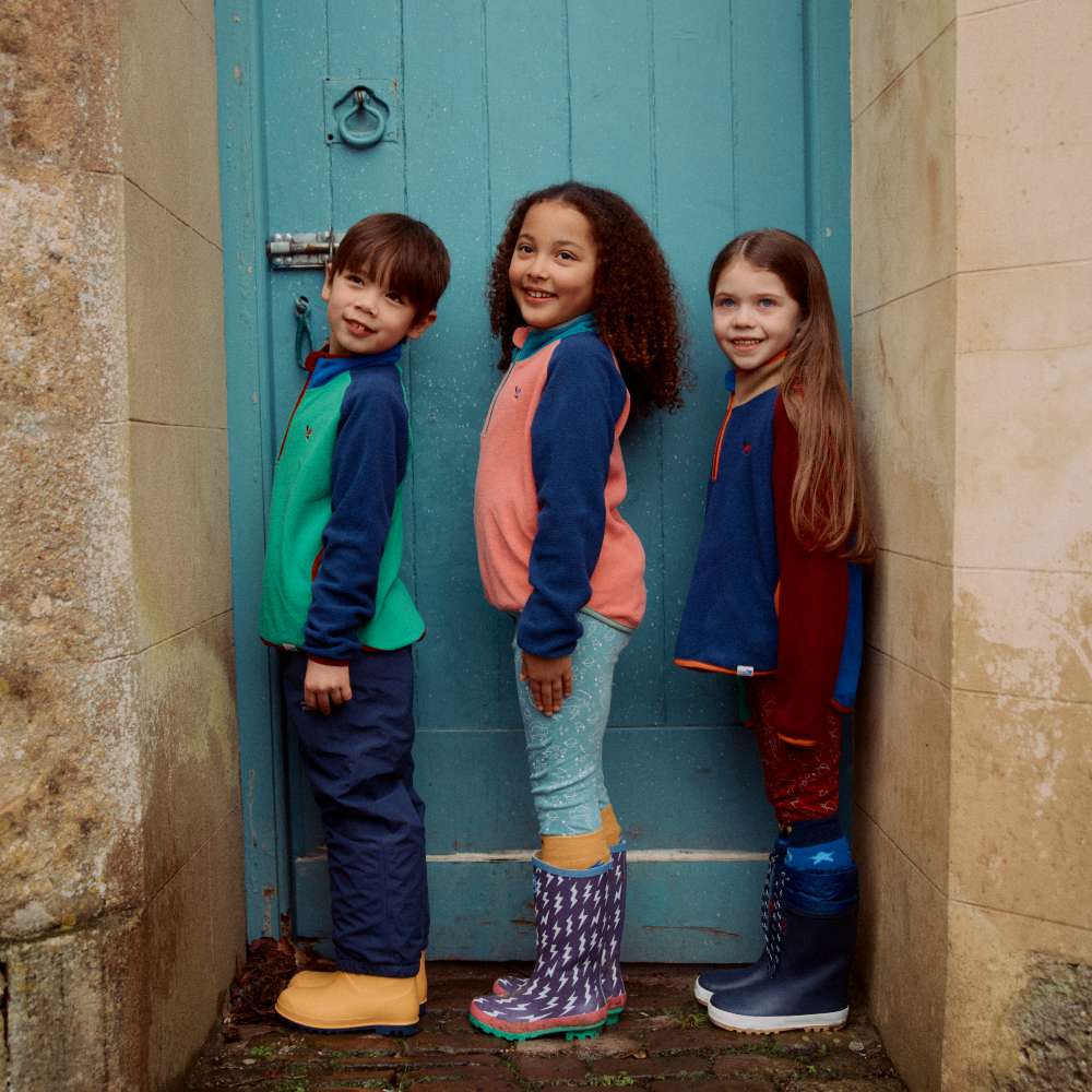 Three children wearing fleeces, trousers and wellies stood outside in front of a blue door 