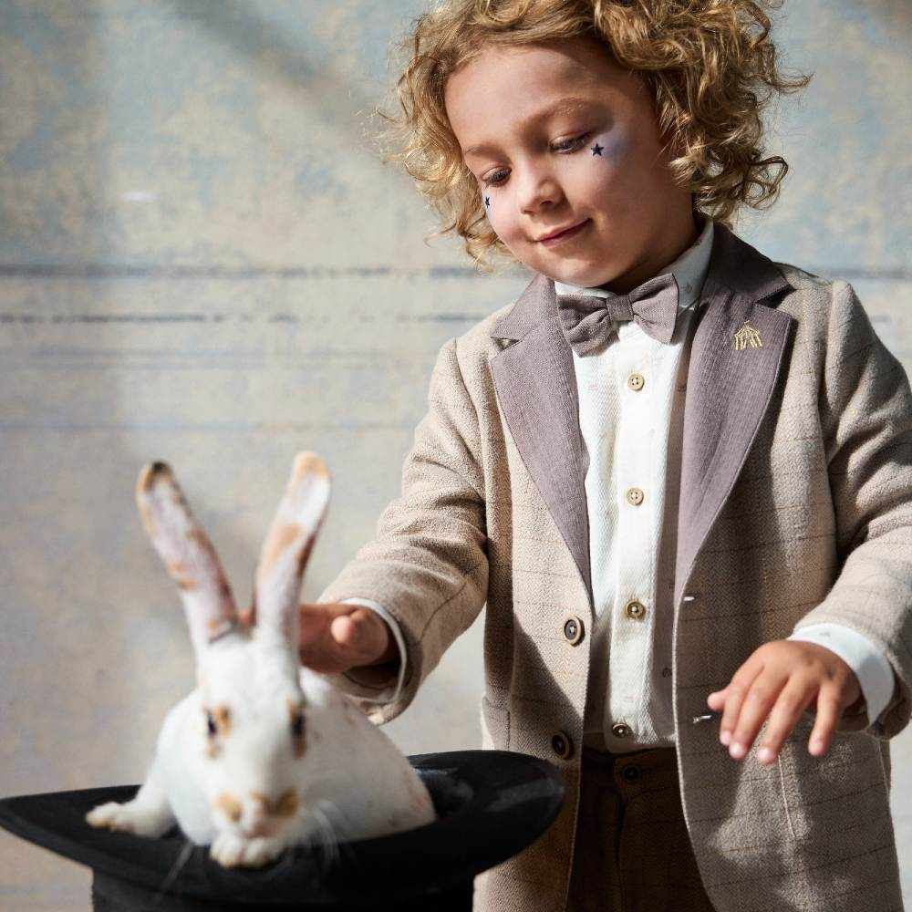 A young child in a blazer, shirt and bowtie stroking a rabbit in a top hat 