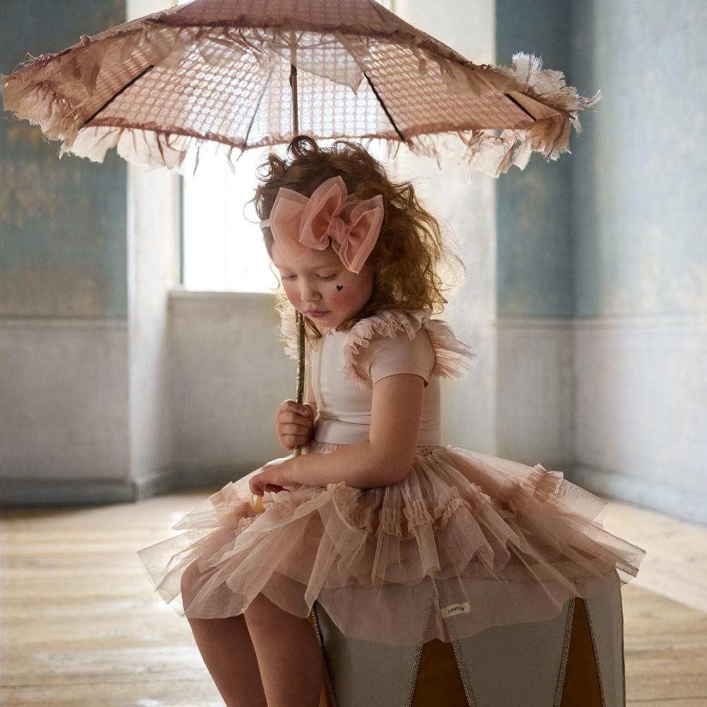 A young girl sat on a circus podium holding a pink parasol and wearing a pink ballerina style outfit