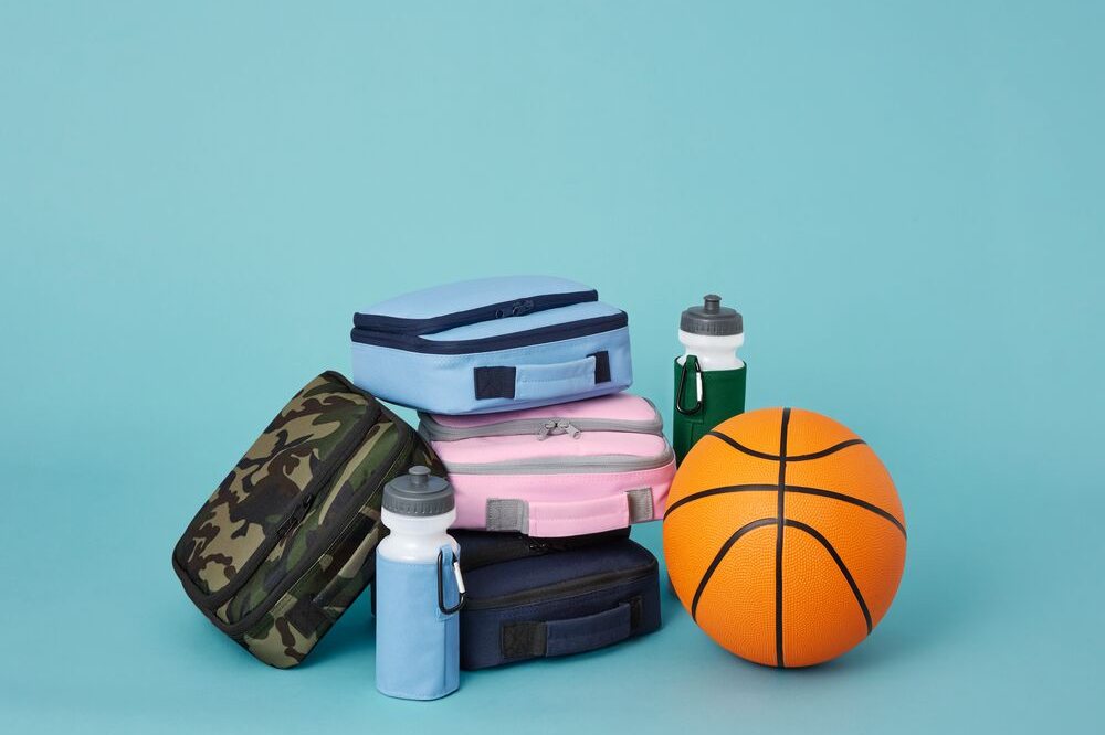 Children's lunch bags and water bottles from the Quadra School Essentials range displayed on a blue background beside a ball