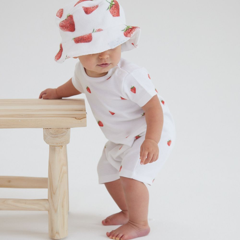 A young child holding onto a chair wearing a hat, top and shorts with a strawberry print on them