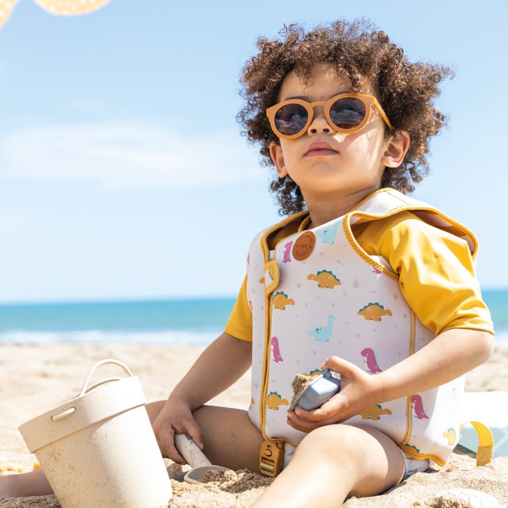 A young child inn a floatsuit and sunglasses playing in the sand on a beach 