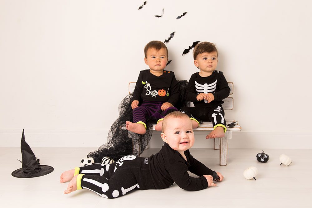 Two young boys sat on a wooden bench with a baby laid on the floor all wearing halloween outfits