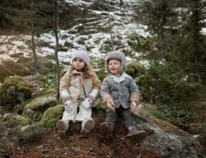 Two children sat on a log in a wood wearing hats and gloves from the Elodie Fairytale Forest collection