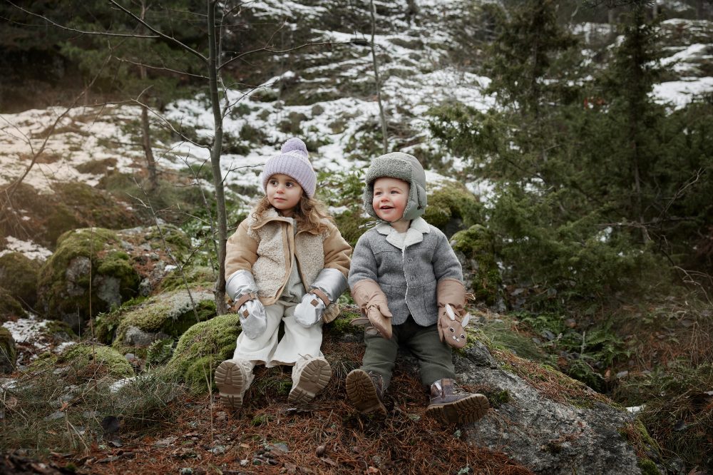 Two children sat on a log in a wood wearing hats and gloves from the Elodie Fairytale Forest collection