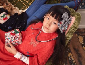 A young girl lying on a sofa looking up at the camera wearing a hairbow and holding a bag