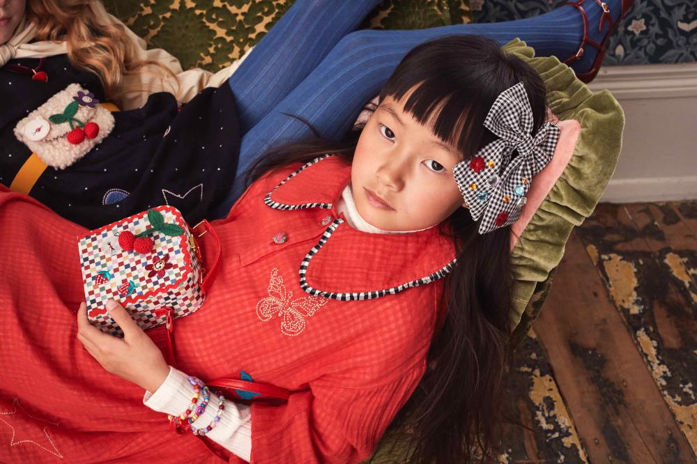 A young girl lying on a sofa looking up at the camera wearing a hairbow and holding a bag