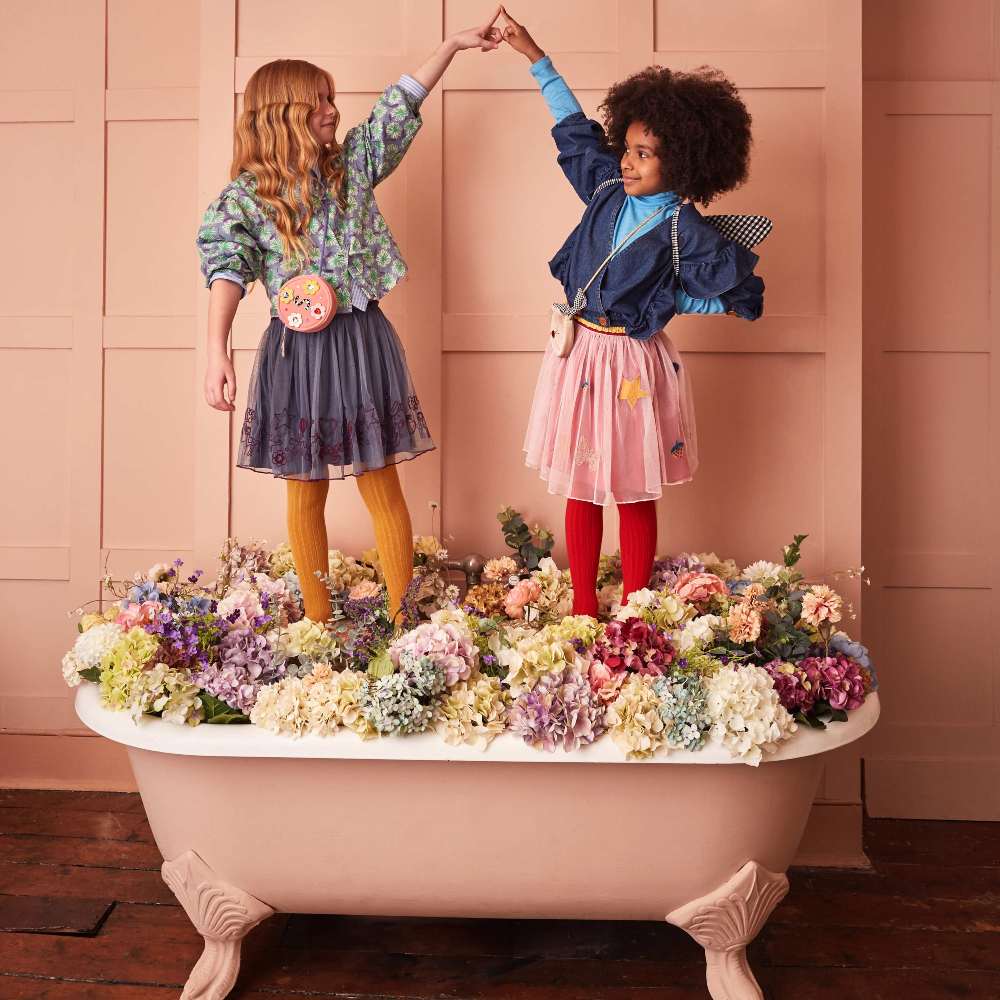 Two girls in a pink room stood in a bath full of flowers raising their hands together 