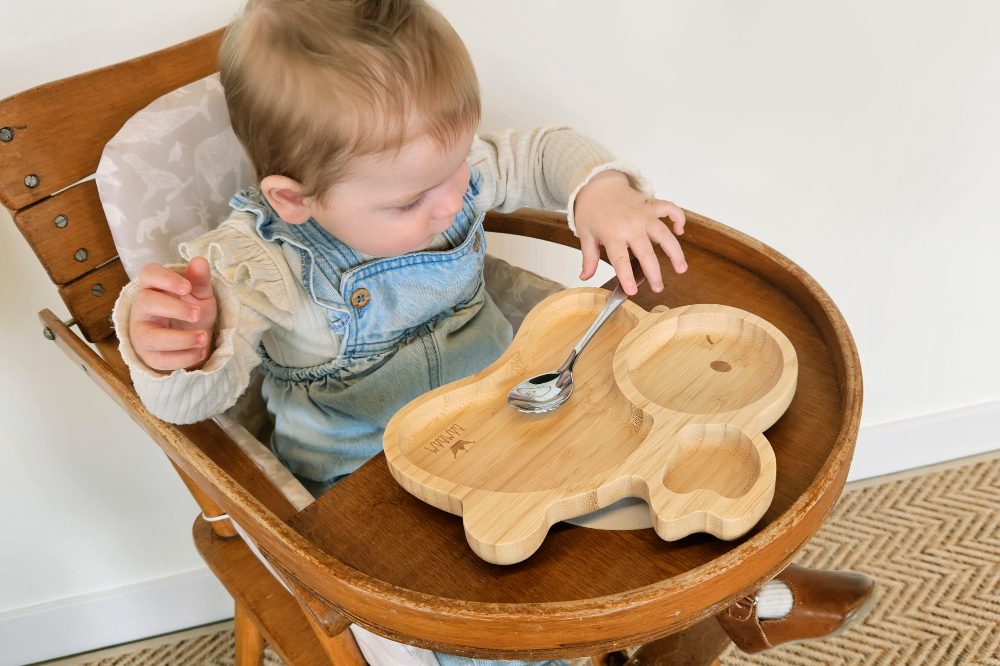 A young child sat in a wooden high chair holding a spoon in front of a wooden rabbit shaped plate