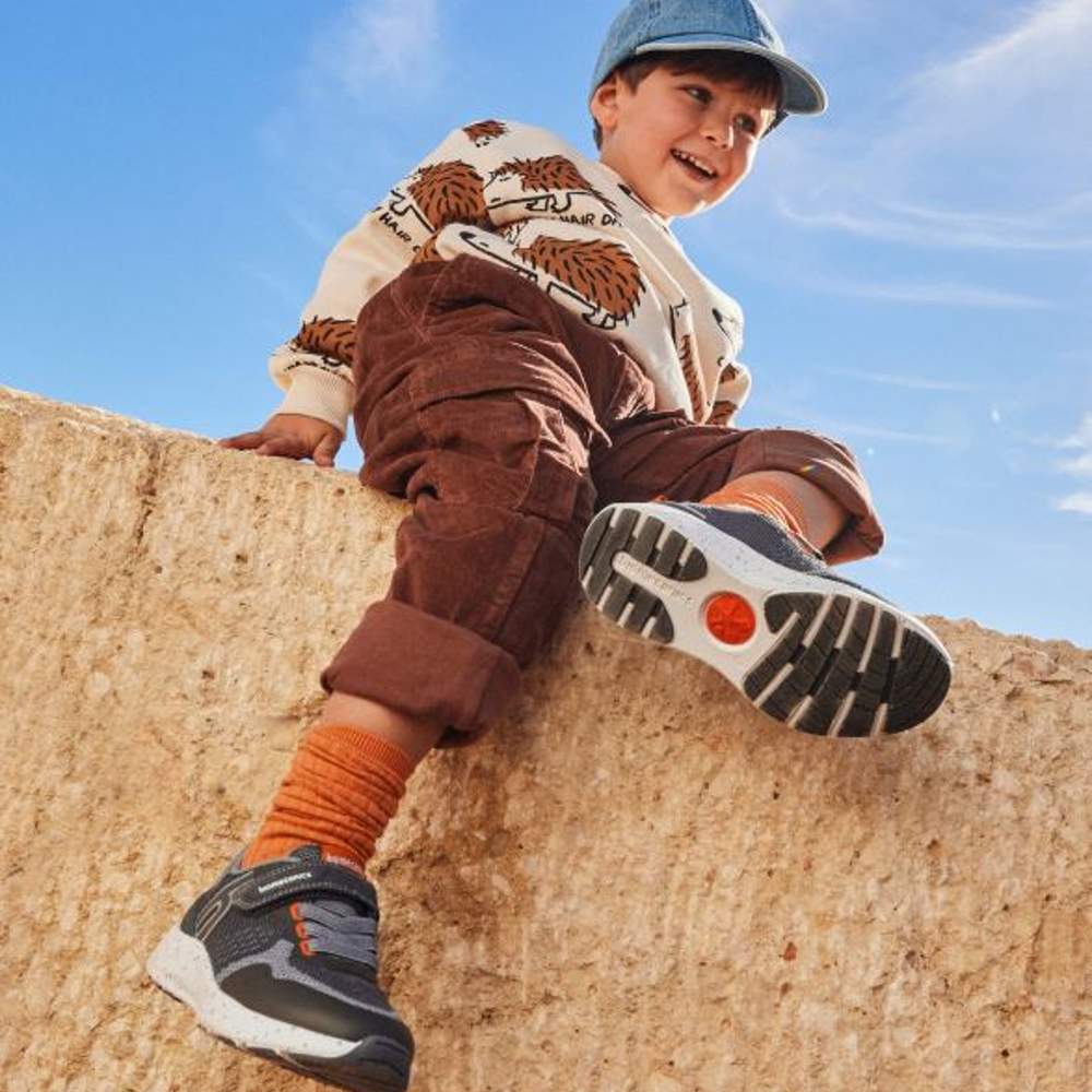 A young boy sat on a wall wearing a cap, top, trousers and trainers 