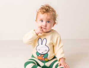 A young child sat on the floor holding their finger to their mouth wearing a top with Miffy on the front and green striped leggings