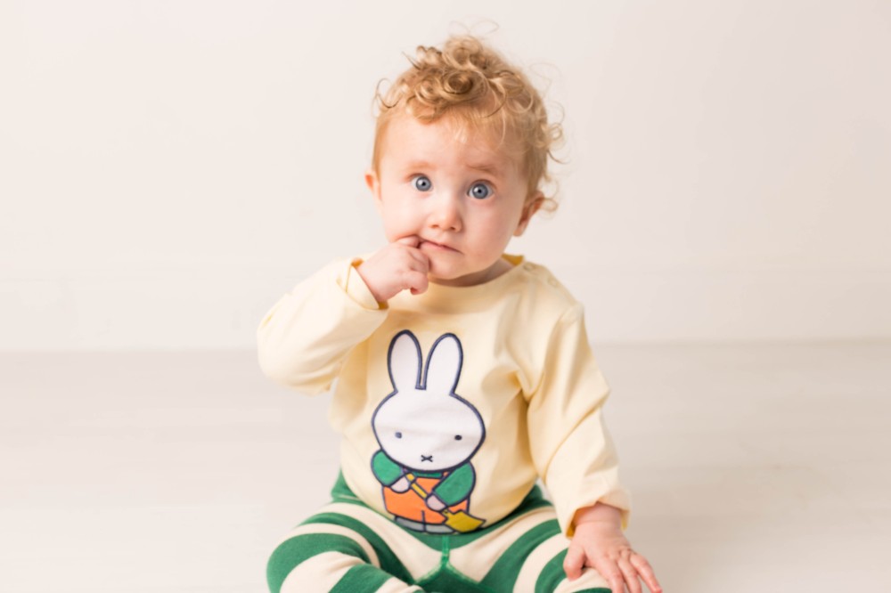 A young child sat on the floor holding their finger to their mouth wearing a top with Miffy on the front and green striped leggings