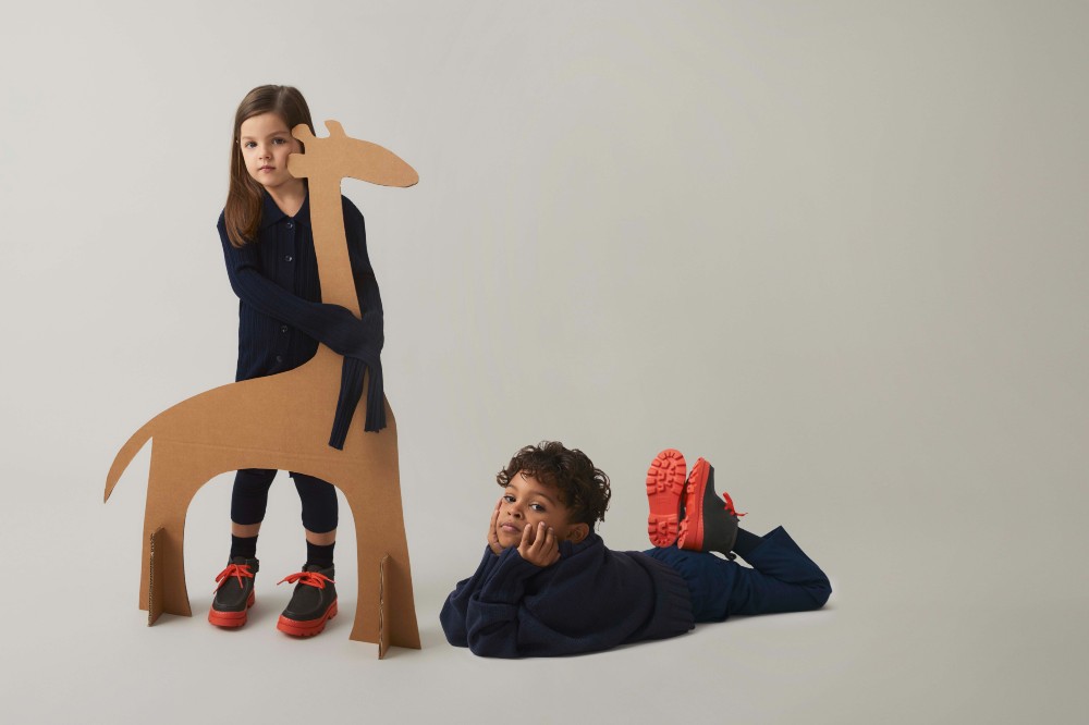 A girl holding a cardboard giraffe beside a boy lying on the floor with his legs in the air 