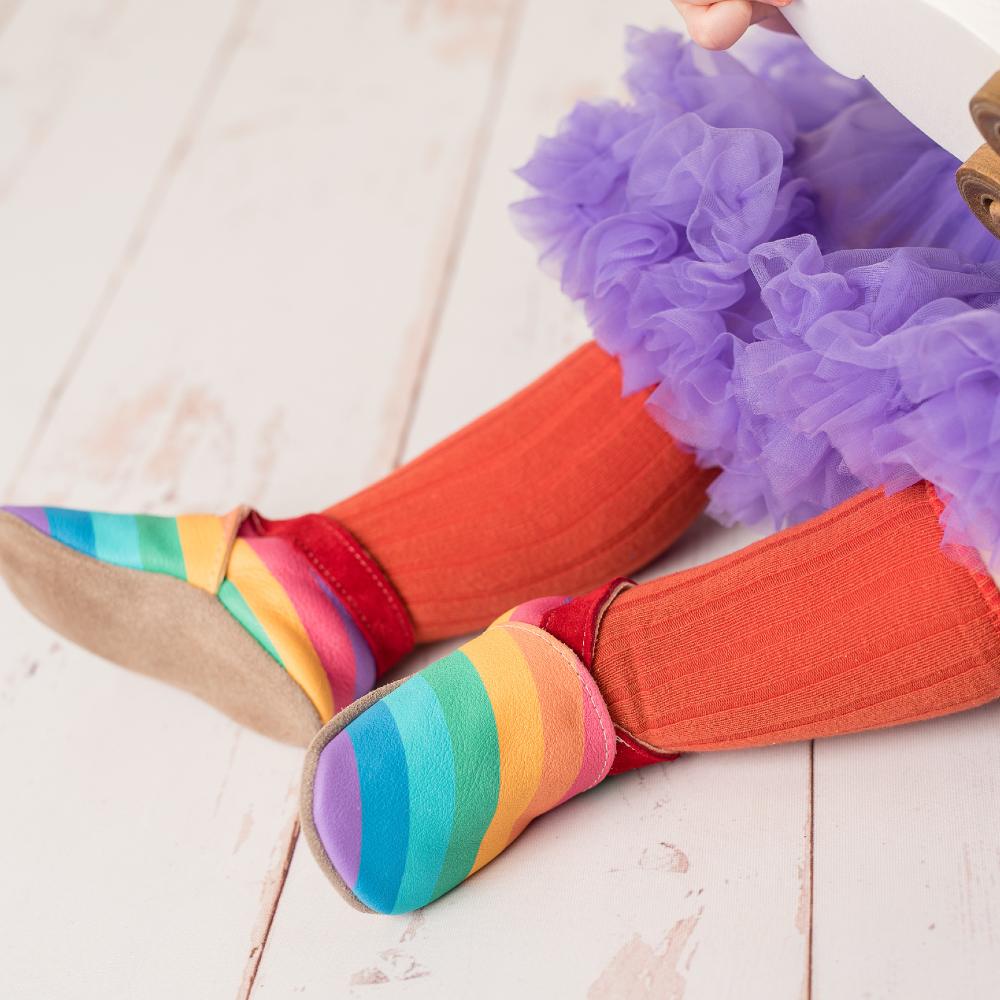 A child's legs shown sat down wearing rainbow soft soled shoes, orange tights and a purple tutu skirt 