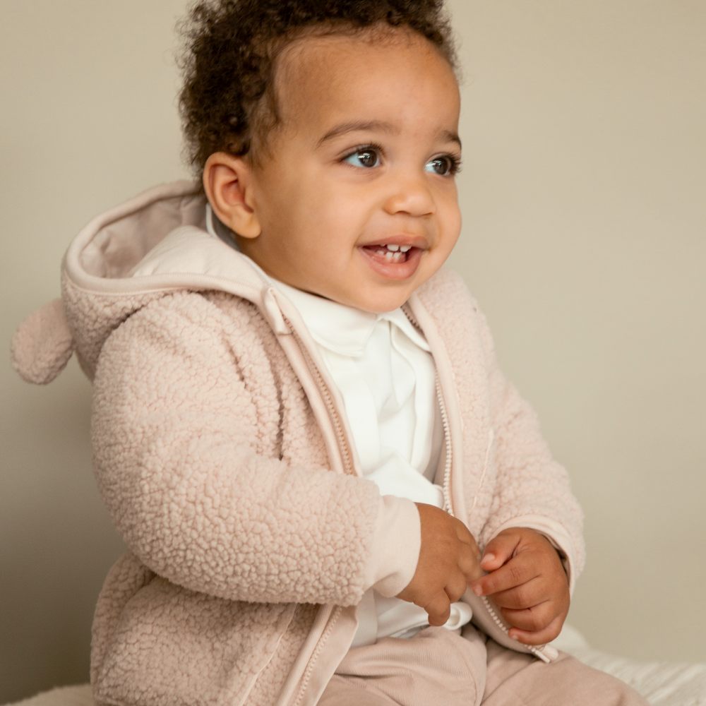 A young child smiling sat down wearing a fleecy jacket and trousers 