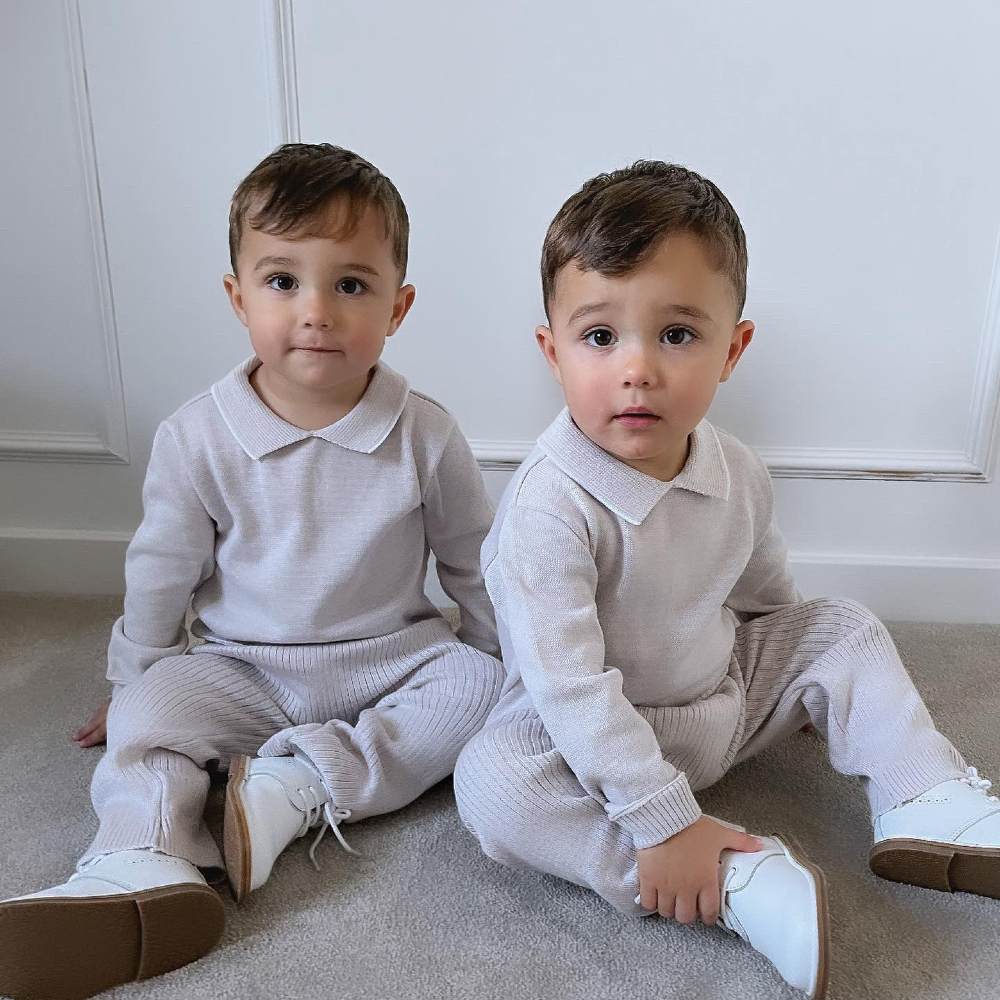 Two young boys sat on the floor wearing matching cream outfits and white shoes 