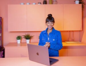 Poppy Child smiling stood in a pink kitchen in front of an open pink laptop to promote The Village App