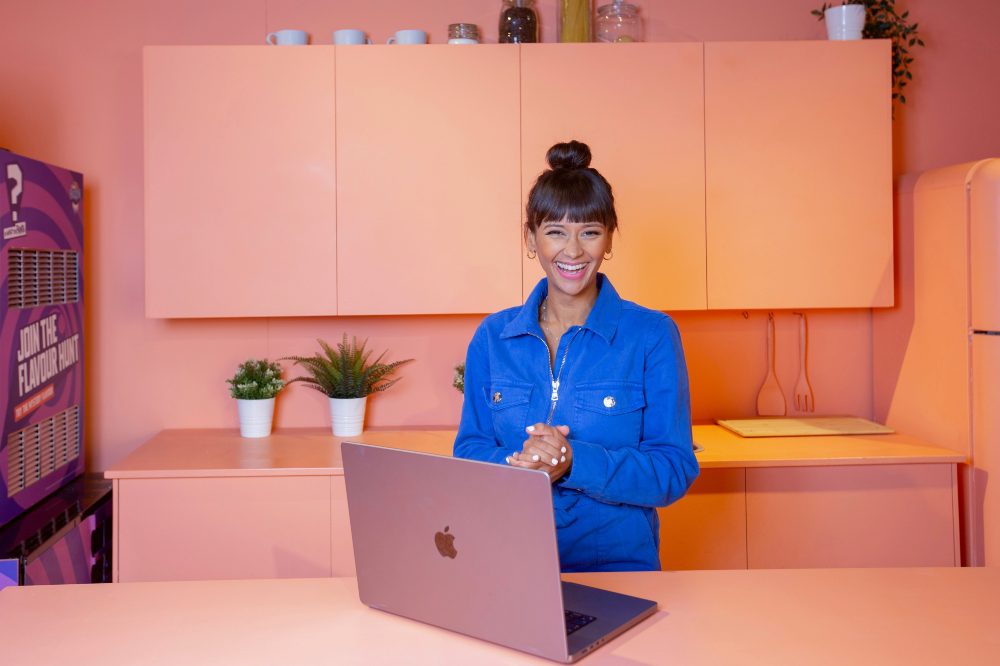 Poppy Child smiling stood in a pink kitchen in front of an open pink laptop to promote The Village App