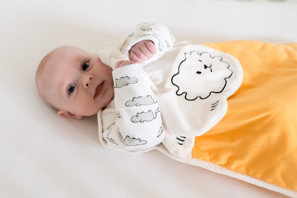 A baby lying down in a hippo print babygro and a yellow sleeping bag by Pure Earth Collection