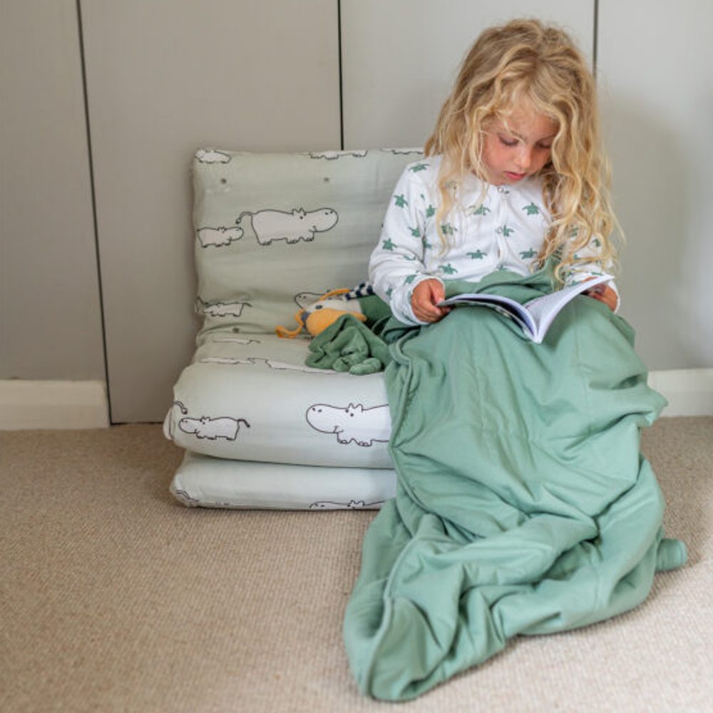 A young girl sat on a fabric chair with a green sleeping bag over her legs reading a book 