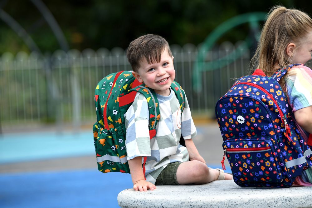 Two young children sat in a playground wearing school rucksacks by Roamie