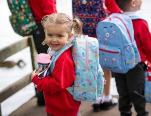 Young school children stood outside with school bags and water bottles by Roamie