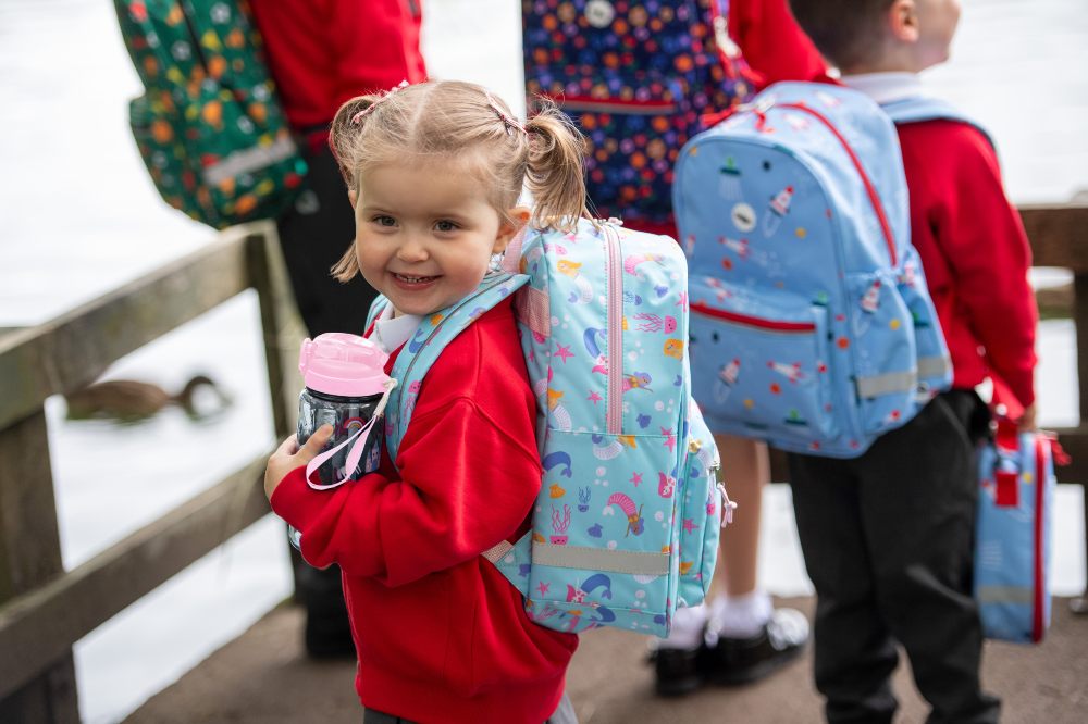 Young school children stood outside with school bags and water bottles by Roamie