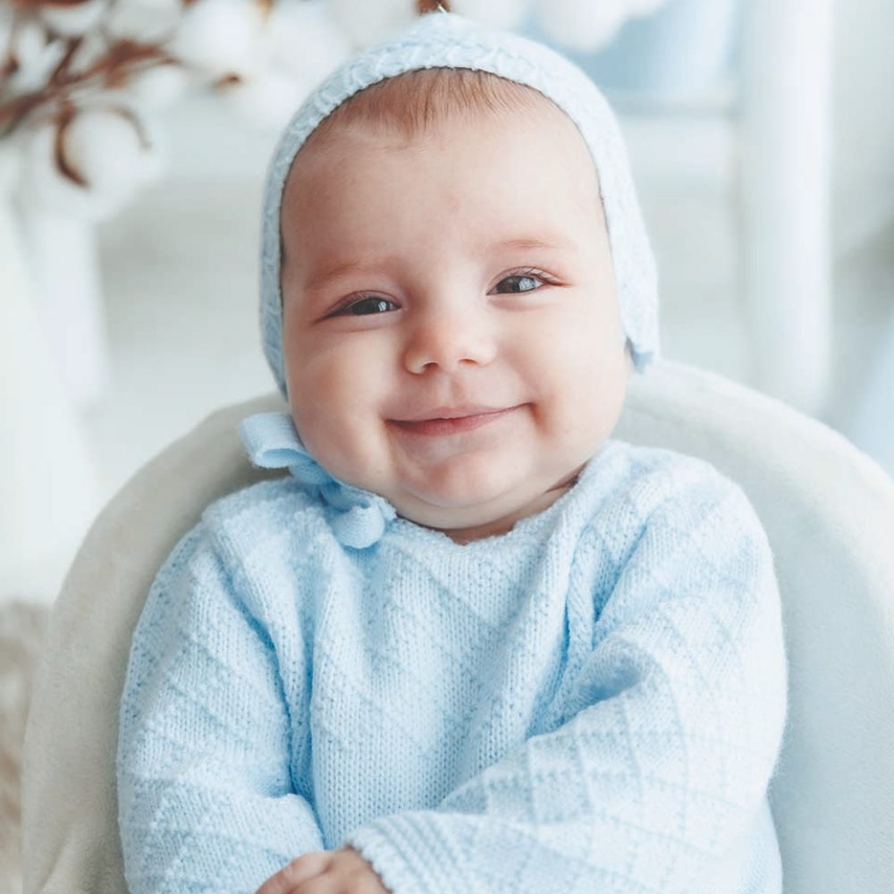 A young baby smiling at the camera wearing a pale blue bonnet and knitted top 