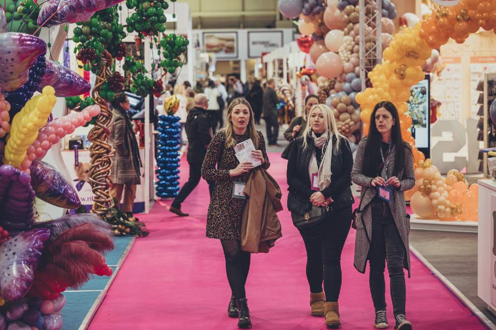 People walking down an aisle at Spring Fair with balloons displayed on stands 