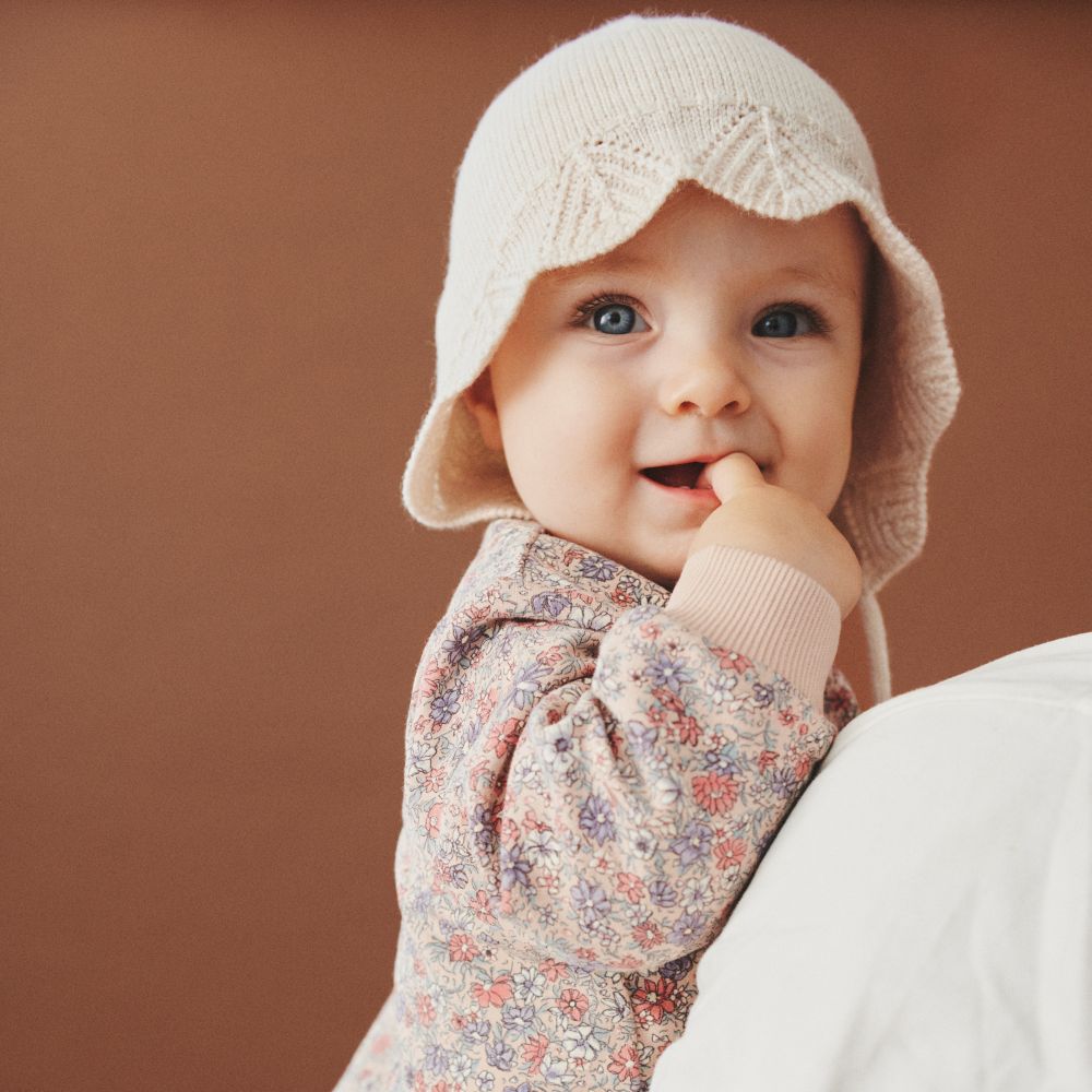 A young baby with their finger in their mouth wearing a crochet hat and floral babygro and being held in someone's arm