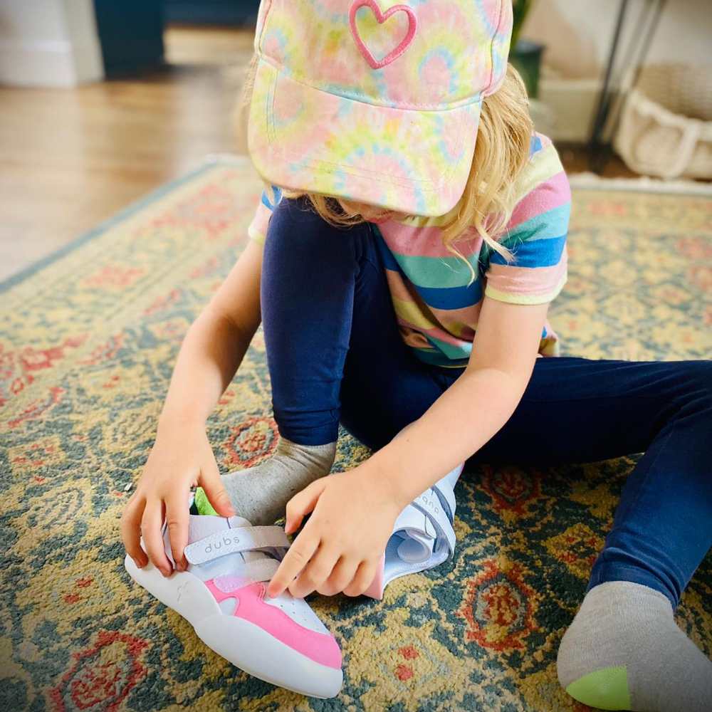 A child sat on the floor wearing a cap putting on a pink and white trainer