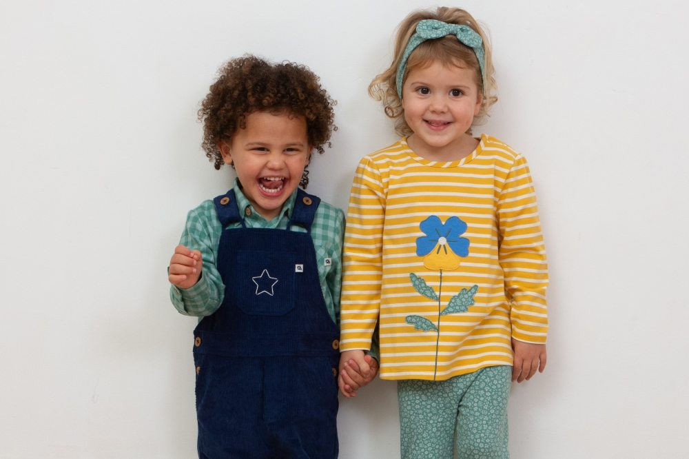 Two young girls holding hands in front of a white wall wearing outfits by the eco-friendly brand Kite Clothing