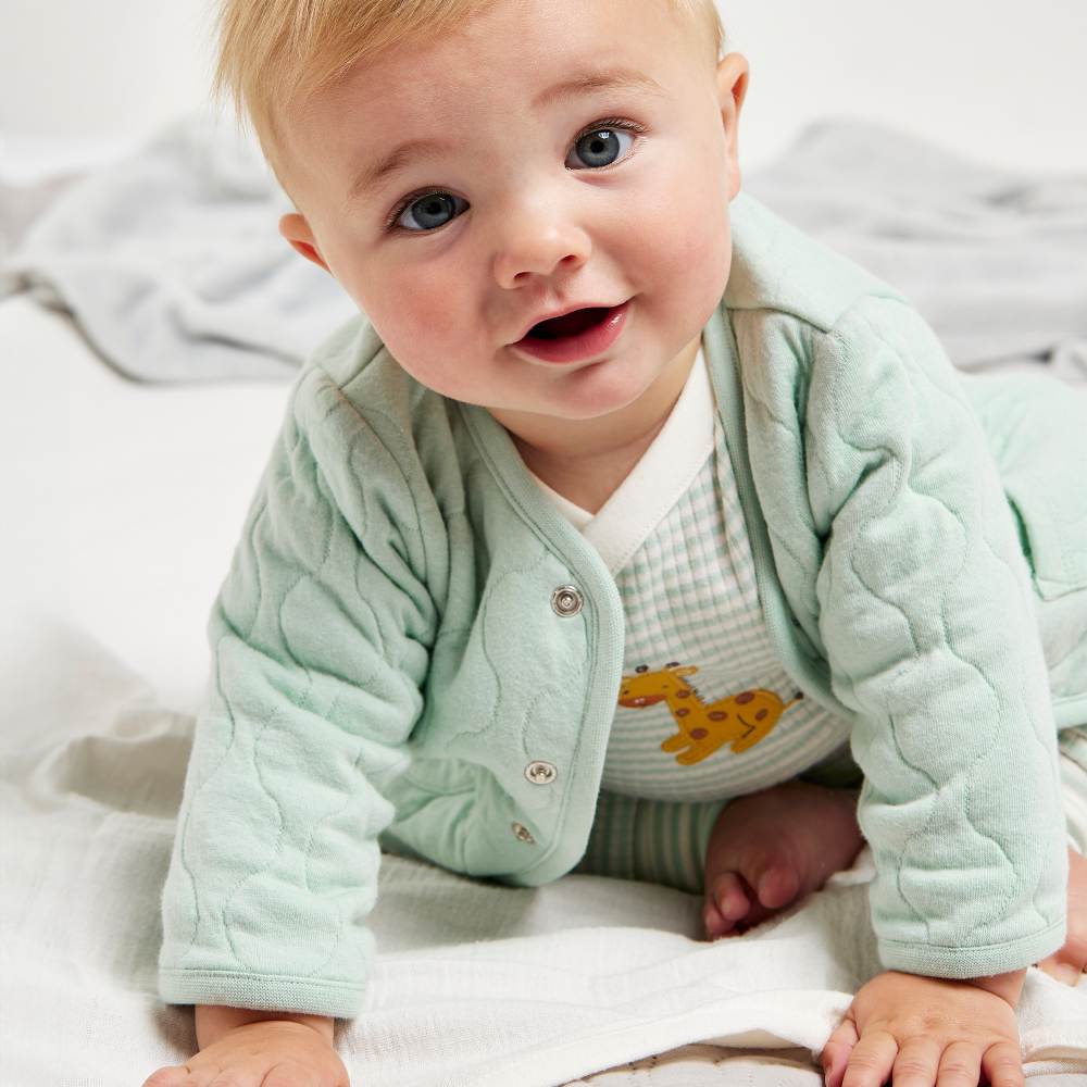 A baby crawling towards the camera wearing a sage green quilted jacket and a top with a giraffe on