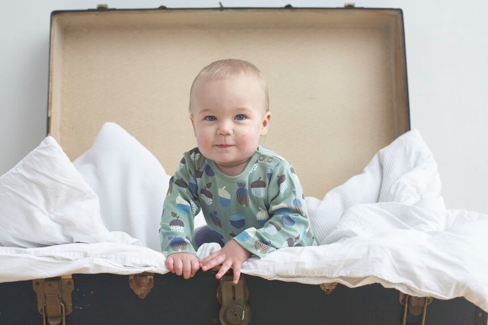 A baby in a vintage suitcase wearing an acorn print babygro by Pigeon Organics