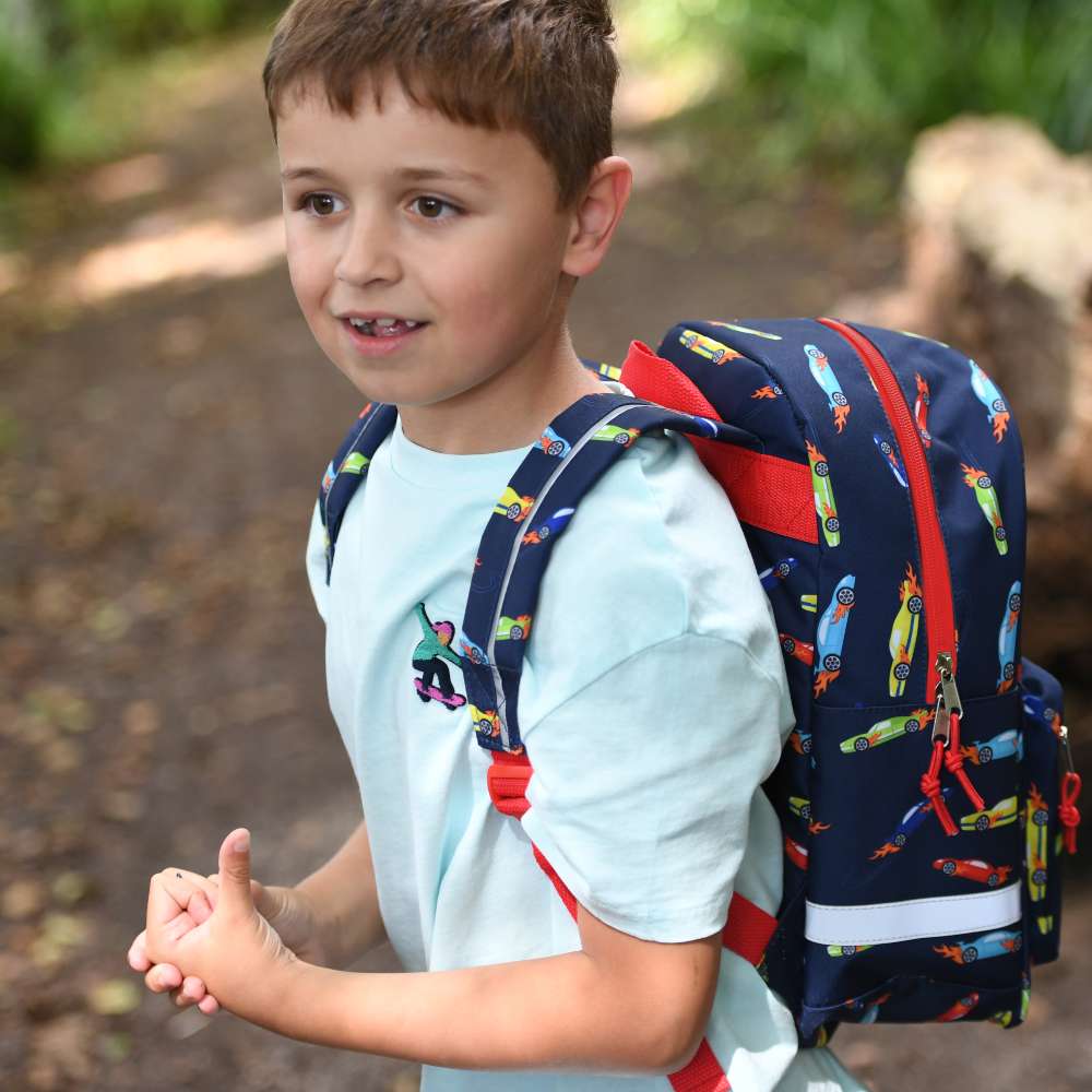 A boy stood outside wearing a blue rucksack with cars on it 