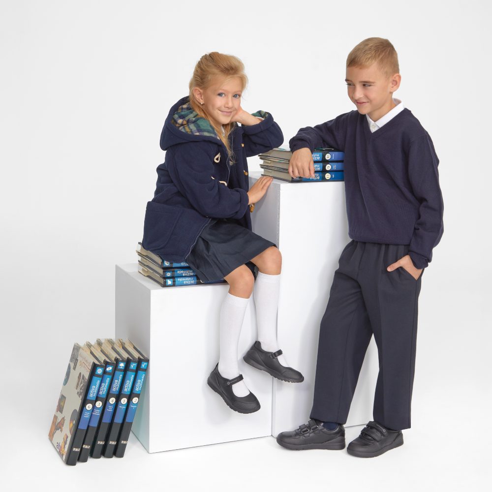 Two children in school uniform sat and stood beside two white pillars and a piles of books 