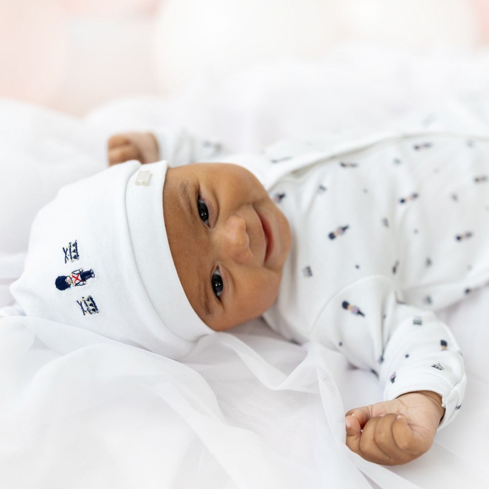 A baby lying on its back wearing a white babygro and hat with a soldier print on them 