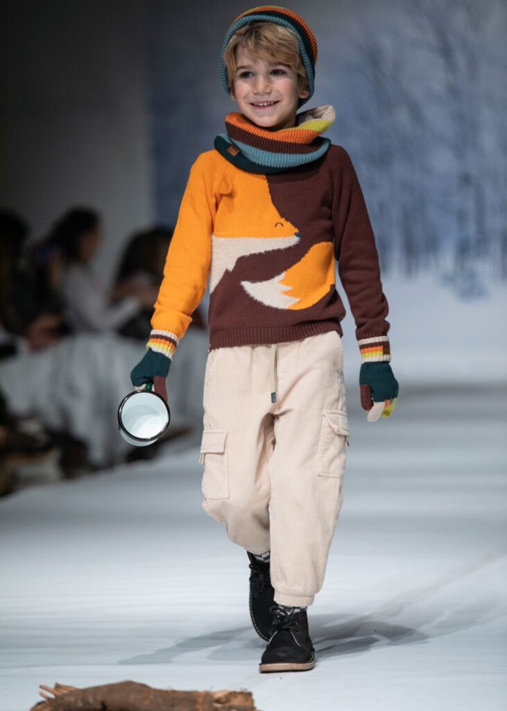 A young boy in a knitted jumper with a fox on it and beige trousers walking down a catwalk 