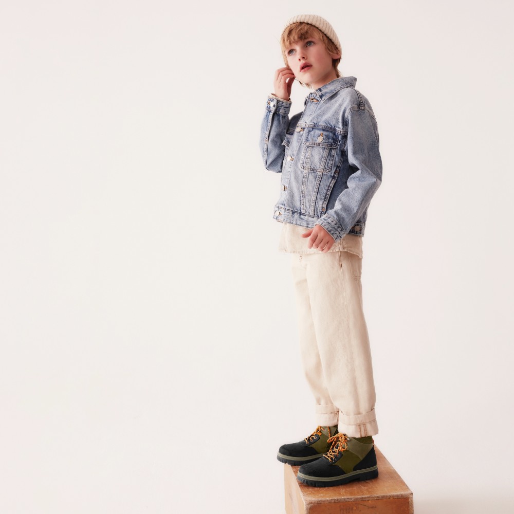 A young boy in a hat, denim jacket, white trainers and boots stood on a wooden box against a white background 