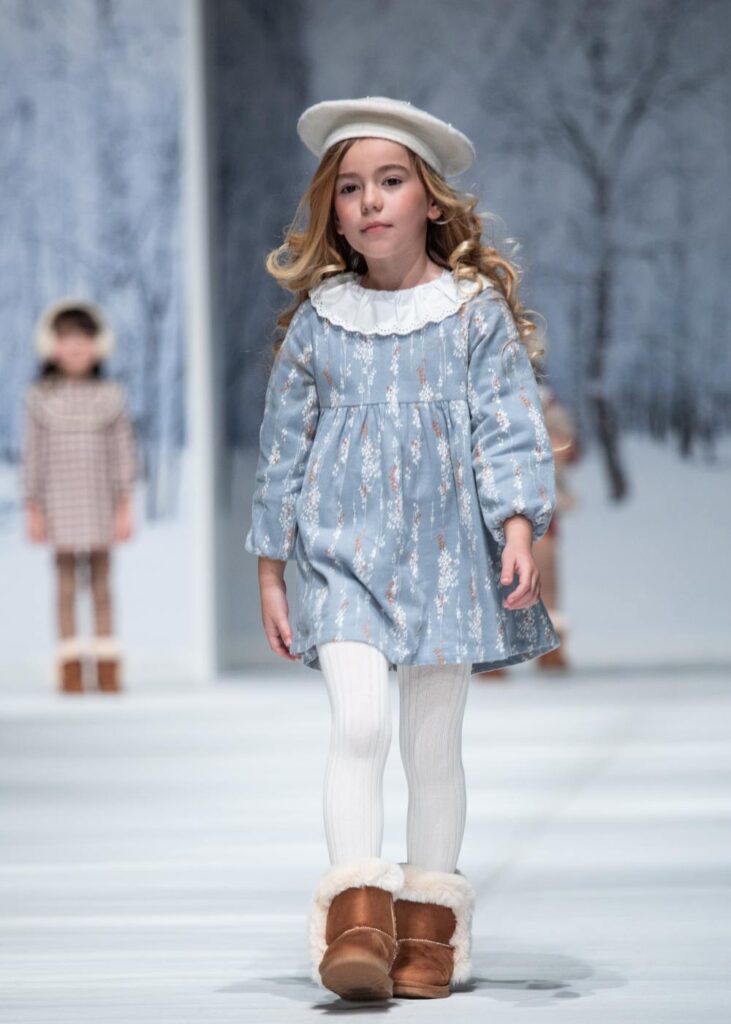 A young girl in a beret, collared dress, white tights and brown boots walking down a catwalk 