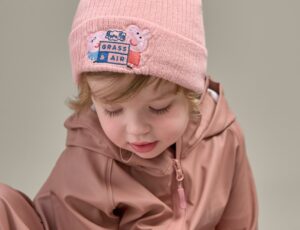 A child in a pink rain jacket and beanie hat with Peppa Pig on the front by Grass & Air