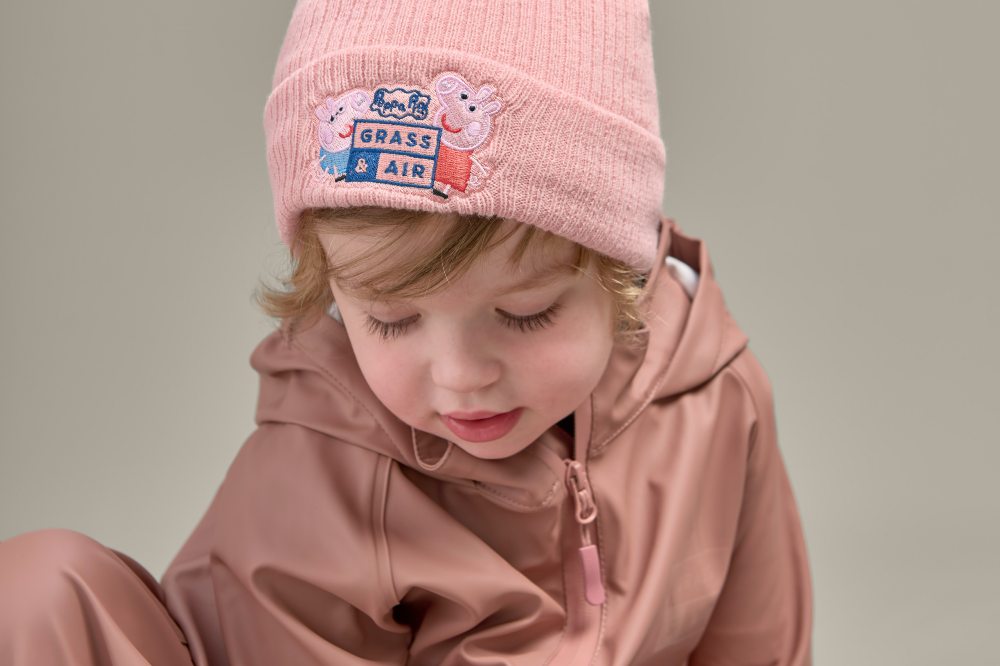 A child in a pink rain jacket and beanie hat with Peppa Pig on the front by Grass & Air