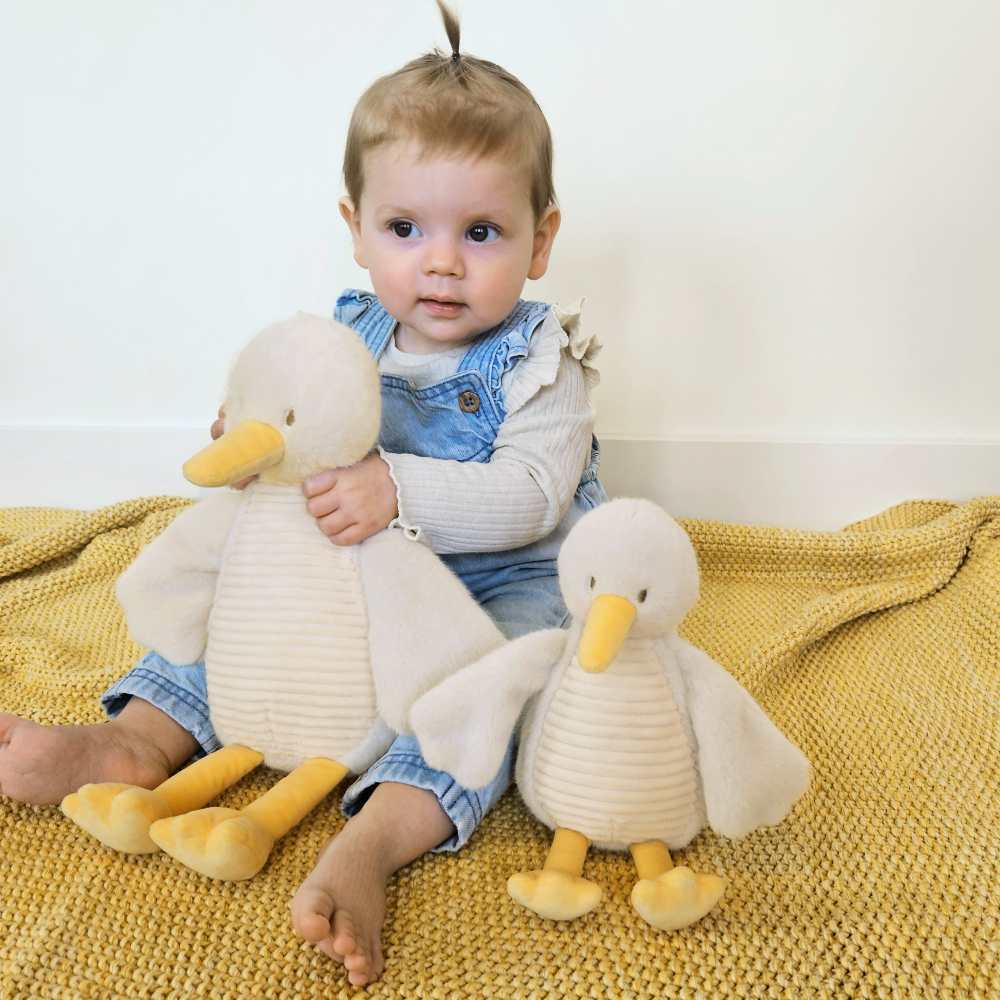 A baby sat on a yellow blanket with two geese soft toys 