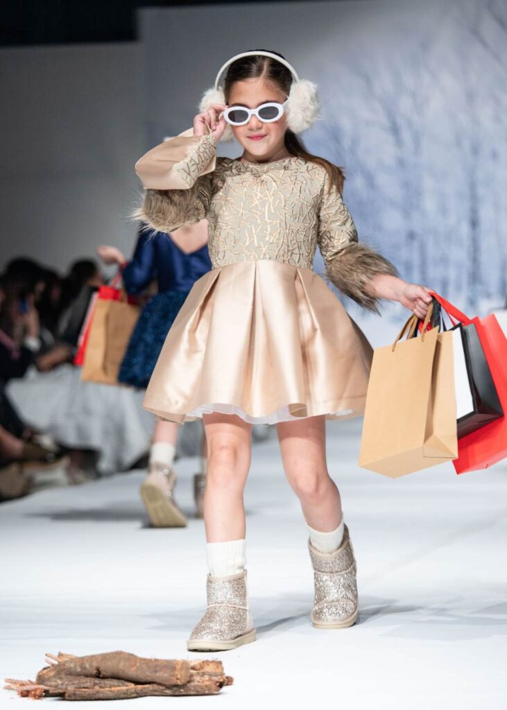 A girl in a gold dress, hat and sunglasses holding shopping bags on a catwalk 