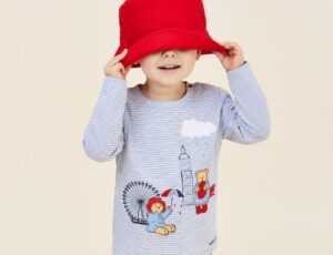 A child pulling a red hat over their eyes wearing a top with Padding Bear on from the JoJo x Paddington collection